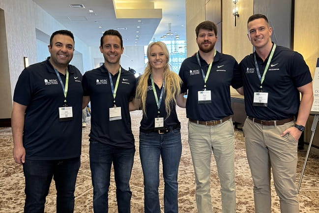 Five residents sporting conference badges and matching MUSC polos stand arm in arm in a conference center lobby.