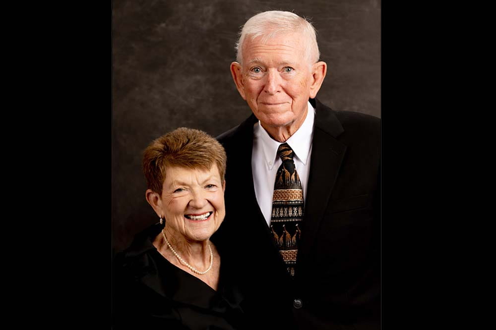 Studio portrait of a tall man wearing a suit and a woman with short reddish brown hair wearing a black top. Both are smiling.